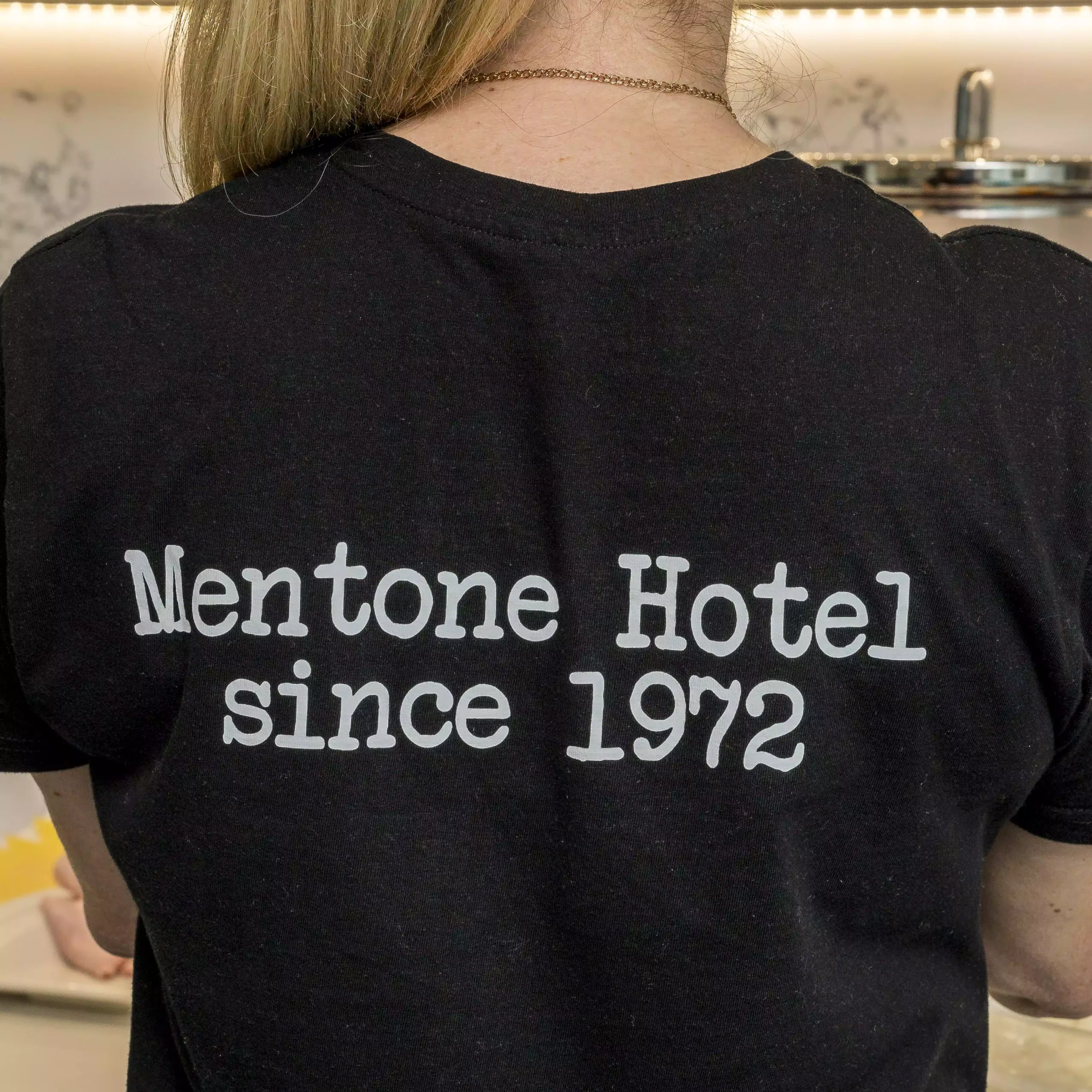 Receptionist in a black shirt with „Mentone Hotel since 1972” printed on the back